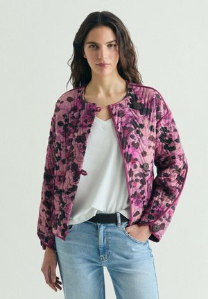 Mujer con una chaqueta acolchada de flores rosa y negra sobre una camiseta blanca con cuello en V y jeans azul claro, de pie con una mano en el bolsillo.