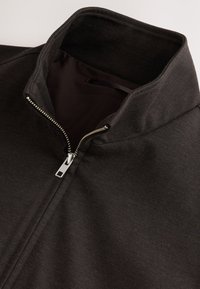 Veste noire avec une fermeture éclair argentée partiellement ouverte et un col haut, montrant la texture lisse du tissu et la doublure intérieure.