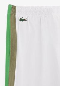 Weiße Shorts mit grünen und braunen Seitenstreifen, aus atmungsaktivem Material, mit einem Logolabel eines Krokodils auf der Vorderseite.