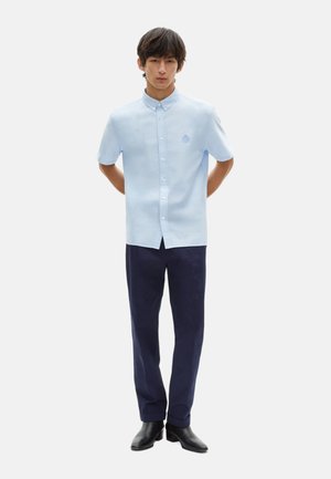 Camisa de botones de manga corta color azul claro con cuello, que presenta un pequeño logotipo bordado, combinada con pantalones oscuros de color navy y zapatos negros.