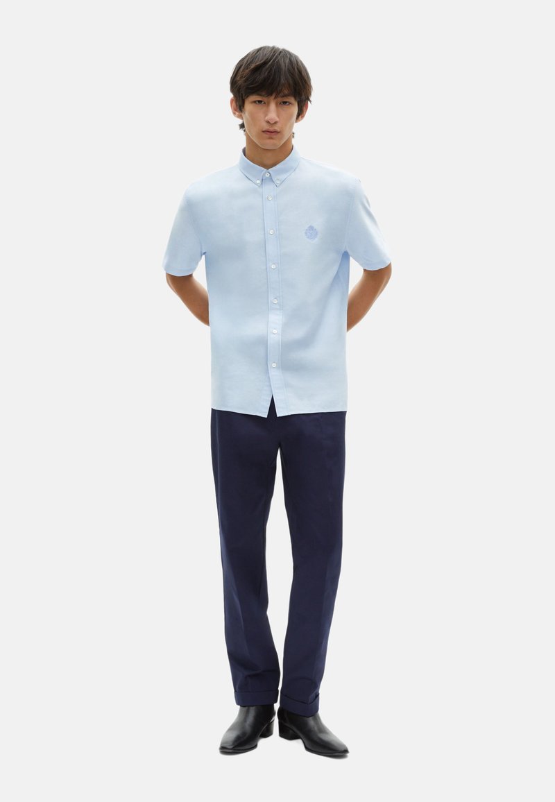 Camisa de botones de manga corta color azul claro con cuello, que presenta un pequeño logotipo bordado, combinada con pantalones oscuros de color navy y zapatos negros.