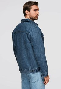 Giacca in denim blu scuro con design classico, caratterizzata da un colletto a punta, tasche laterali e chiusura a bottoni a pressione all'orlo.