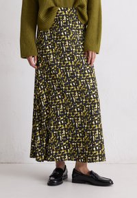 Maxi-jurk in een gedurfde zwarte, gele en witte geometrische print, gemaakt van een gladde stof. Gecombineerd met een gestructureerde groene trui en zwarte loafers.
