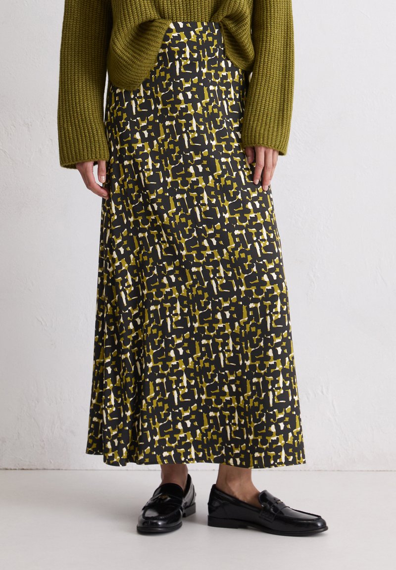 Maxi-jurk in een gedurfde zwarte, gele en witte geometrische print, gemaakt van een gladde stof. Gecombineerd met een gestructureerde groene trui en zwarte loafers.