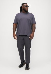 Graues Baumwoll-T-Shirt mit Rundhalsausschnitt und einem kleinen gelben Logo auf der Brust, kombiniert mit dunkelgrauen Cargo-Hosen und schwarzen Schuhen.