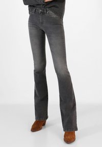 Gray flared jeans in denim with a distressed finish. Features a mid-rise waist, five pockets, and stitching details. Paired with brown ankle boots.