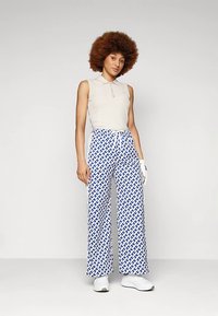 Beige ärmlös pikétröja kombinerad med vida byxor med ett blått och vitt geometriskt mönster, kompletterat med vita skor.