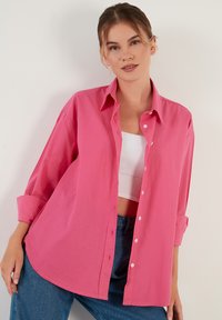 Pink button-up shirt made of lightweight cotton, featuring a collar, long sleeves with rolled cuffs, and a curved hem. Simple, solid color design.