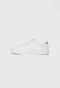 Lage witte sneaker met gouden accenten, gestructureerde bovenkant, witte veters en "Tommy Hilfiger" branding aan de zijkant.