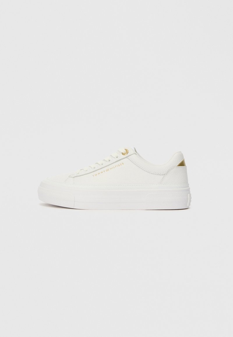 Lage witte sneaker met gouden accenten, gestructureerde bovenkant, witte veters en "Tommy Hilfiger" branding aan de zijkant.
