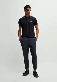 Jūras zila polo krekls ar baltajiem akcentiem, kuram ir jaunums. Apvienots ar šauriem jūras zila biksēm un melniem sporta apaviem. Brilles pabeidz komplektu.