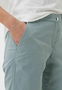 Lichtblauwe broek met zijzakken, gemaakt van een gladde stof. Een zilveren armband met een ronde accent wordt om de pols gedragen.