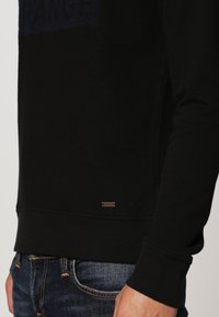 Sudadera negra con una textura suave, que presenta un sutil detalle del logo en el dobladillo y un texto azul en contraste en el pecho.