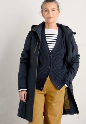 Navy waterdichte trenchcoat met een capuchon, gelaagd over een donkere gebreide vest en een gestreept shirt, in combinatie met mosterdkleurige broeken.
