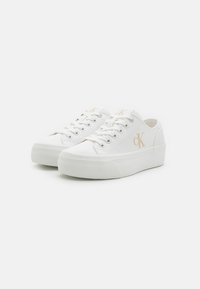 Baskets en toile blanche avec une semelle compensée, dotées de lacets plats, d'un logo discret sur le côté et d'une texture lisse.