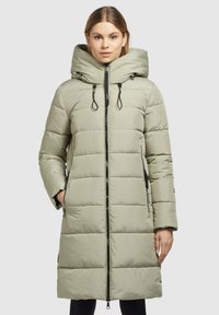 Mujer con abrigo largo acolchado en color beige claro y cuello alto, con detalle de cremallera negra, de pie con una mano en el bolsillo frente a un fondo liso.