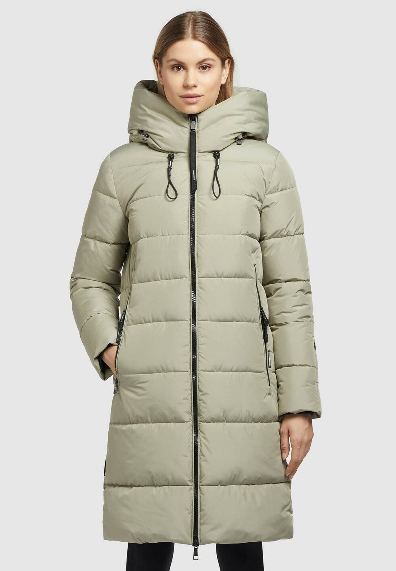 Mujer con abrigo largo acolchado en color beige claro y cuello alto, con detalle de cremallera negra, de pie con una mano en el bolsillo frente a un fondo liso.