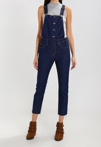 Woman wearing dark blue denim overalls with buttons, a sleeveless gray turtleneck top, and brown ankle boots with buckles.