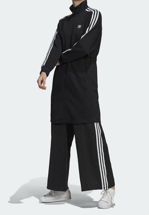 Person wearing black Adidas long zip jacket and wide-legged pants with white stripes, standing with arms crossed.