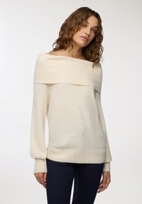 Maglione crema off-shoulder con colletto e polsini a costine. Tessuto morbido, maniche lunghe e vestibilità ampia. Abbinato a jeans scuri.