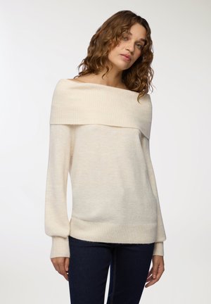Maglione crema off-shoulder con colletto e polsini a costine. Tessuto morbido, maniche lunghe e vestibilità ampia. Abbinato a jeans scuri.