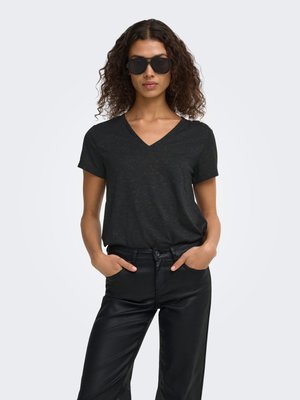 Vrouw met krullend haar die donkere zonnebril draagt, een zwart T-shirt met V-hals en korte mouwen, en zwarte broek met handen in de zakken.