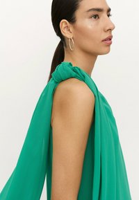 Top de gasa verde con un diseño drapeado, detalle de hombro anudado y textura suave, con una silueta relajada que destaca un suave flujo.