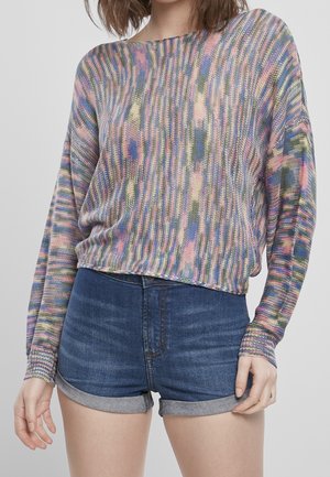 Femme portant un pull en tricot multicolore à manches longues et un short en jean taille haute aux bords retroussés, se tenant devant un fond uni.