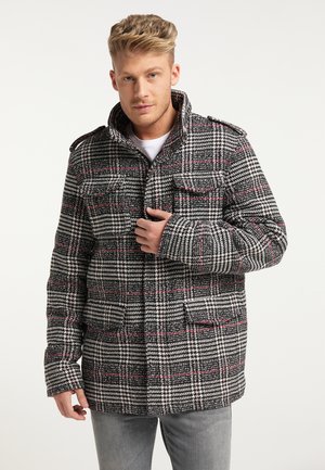 Man in zwart-wit geruit jasje met rode lijnen, draagt lichtgrijze jeans, raakt de voorkant van het jasje bij de kraag aan.