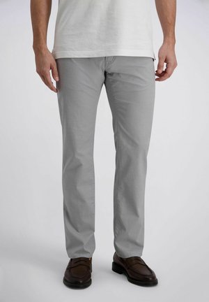 Mann trägt hellgraue gerade geschnittene Hose, braune Lederschuhe und ein weißes Hemd, steht vor einem schlichten Hintergrund.