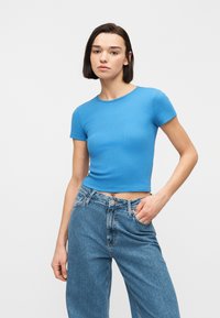 Νέα γυναίκα με κοντά σκούρα μαλλιά, που φοράει στενό κοντό μπλουζάκι σε μπλε απόχρωση και φαρδιά μπλε τζιν, στέκεται μπροστά σε λευκό φόντο.