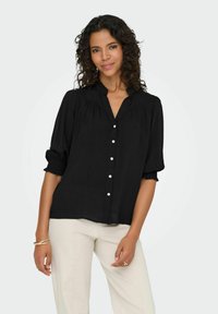 Sort knap-up skjorte med en blød tekstur, korte puffede ærmer, indsamlet detalje ved skuldrene og krave. Sættes sammen med lyse bukser.