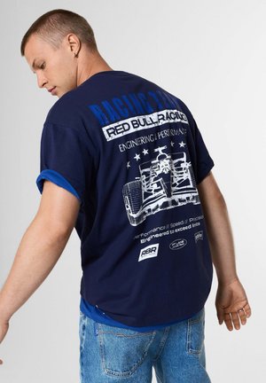 Hombre que lleva una camiseta azul marino de Red Bull Racing con un gráfico blanco de un coche de carreras y texto en la espalda, combinado con vaqueros azules.