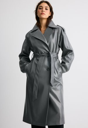 Giovane donna che indossa un trench coat in pelle grigia con cintura e ampi revers, in piedi con le mani nelle tasche contro uno sfondo semplice.