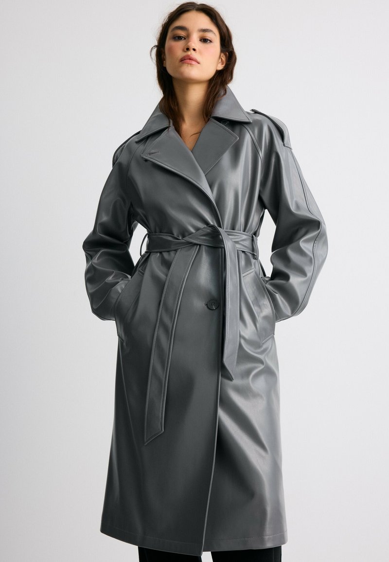 Jeune femme portant un trench-coat en cuir gris ceinturé avec de larges revers, debout les mains dans les poches devant un fond uni.