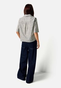 Žena s dlouhými hnědými vlasy, oblečená v krátkém vzorovaném tričku a širokých tmavě modrých džínách, stojící zády k bílému pozadí.