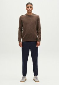 Barna kötött pulóver kerek nyakkivágással, sötétkék nadrággal és világosszürke tornacipővel párosítva. Egyszerű, texturált anyag és bordázott mandzsetták.