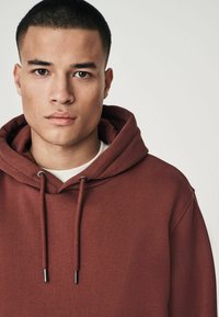 Sudadera con capucha marrón, con cordones, de textura suave y un corte relajado. Camiseta blanca visible debajo de la sudadera.