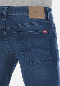 Vaqueros de mezclilla en azul oscuro con un acabado texturizado. Presentan un parche de marca de cuero marrón y una pequeña etiqueta roja en el bolsillo trasero.