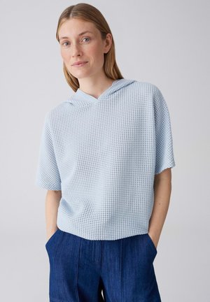 Vrouw met lichtbruin haar, gekleed in een lichtblauwe, gestructureerde korte mouwen hoodie en donkerblauwe broek, met handen in de zakken.