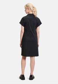 Schwarzes kurzärmeliges Kleid mit Kragen, tailliertem Rücken und geradem Schnitt, kombiniert mit klobigen schwarzen Schuhen, die eine glatte Stofftextur zeigen.
