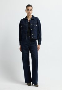 Ciemnoniebieska dżinsowa kurtka z złotymi guzikami, w połączeniu z wysokim stanem ciemnych dżinsów. Oba elementy mają luźny krój. Tekstura jest gładka i wytrzymała.