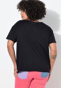 Mujer con cabello rizado de pie de espaldas, vistiendo una camiseta negra suelta y pantalones rosas con bolsillos traseros morados y azules, mostrando tatuajes en los brazos.