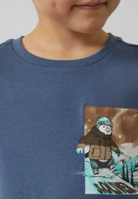 Niebieska bawełniana bluza z nadrukiem pingwina na snowboardzie, ubranego w brązową kurtkę i niebieski kask na tle śnieżnego krajobrazu.