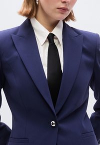 Μπλε σκούρο κομψό σακάκι με ένα ασημένιο κουμπί, πέτα  κορυφής και δομημένη γραμμή, σε συνδυασμό με μαύρη γραβάτα πάνω από λευκό πουκάμισο.