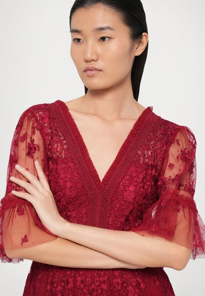 Punane pitskleit sügava V-kaelusega, läbipaistvate tikandiga käiste ja volangakujuliste aktsentidega. Kangas on kaetud tekstuurse lillemustri ning see lisab rõivale ilu.