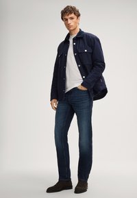 Dunkelblaue Hemdjacke mit zwei Brusttaschen und Druckknöpfen, kombiniert mit blauen Jeans und dunkelbraunen Stiefeletten, vor neutralem Hintergrund.