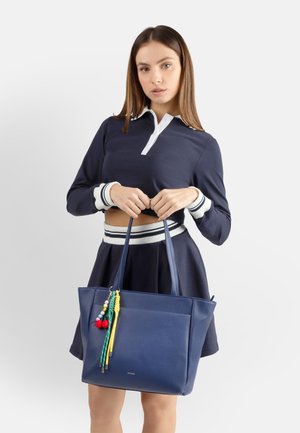 Giovane donna con outfit blu marino che tiene una grande borsa tote blu marino decorata con nappine colorate su uno sfondo semplice.