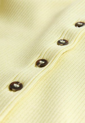 Nahaufnahme eines blassgelben gerippten Stoffes mit einer Reihe dunkeloranger Knöpfe, die in einer vertikalen Linie genäht sind.