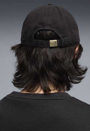 Persoon met schouderlang donker haar, draagt een zwarte Puma-pet achterstevoren en een zwart shirt met ronde hals, gezien van achteren.